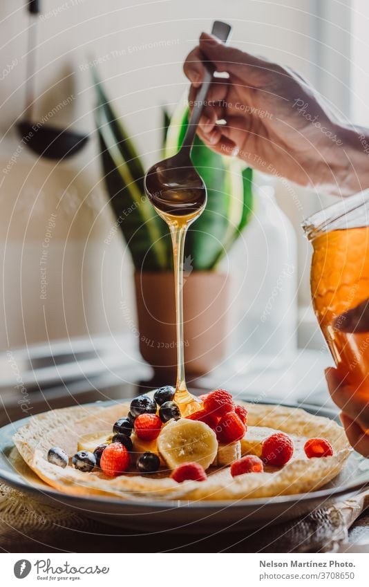 Delicious crepe with bananas, blueberries, raspberries and honey. It is lit with a nice natural light in the kitchen. We can see the honey falling from the spoon.