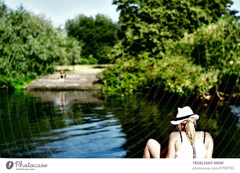The sun shines on the pale skin of bare shoulders and the naked, bent leg of a young woman with a straw hat and blond ponytail, sitting on the quay wall and looking into the water in front of the shore of the tree-covered Friendship Island