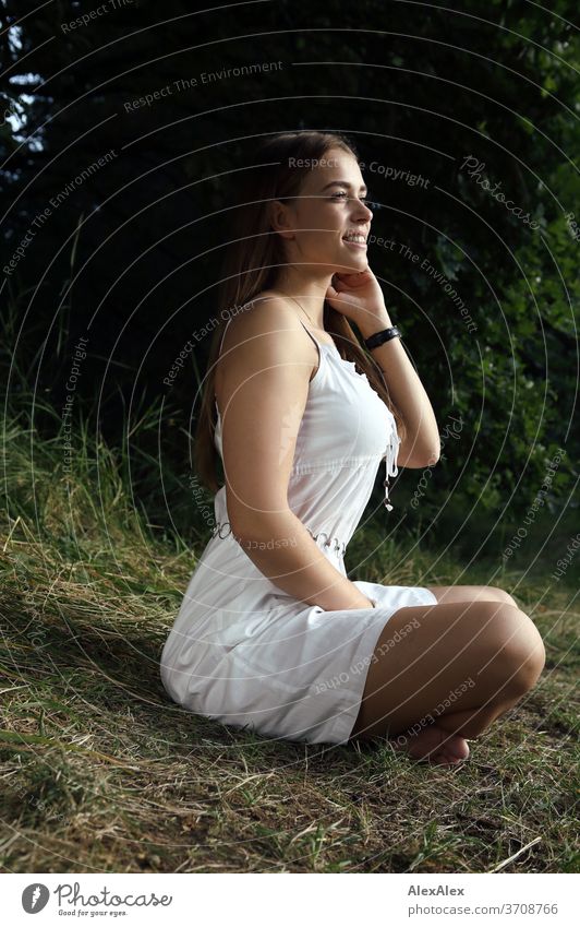 Portrait of a young woman in a white summer dress sitting cross-legged in nature Light Athletic Feminine Emotions emotionally Looking into the camera portrait