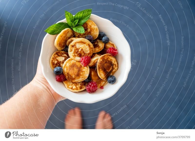 Hand with fresh little pancakes in a white bowl. Fresh berries, top view. Small Berries by hand stop scatterbrained Barefoot plan Blue floor Breakfast Roasted