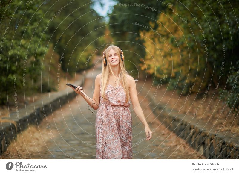 Smiling woman with smartphone and headphones walking in park happy listen using music summer enjoy young female browsing alley path device gadget lifestyle