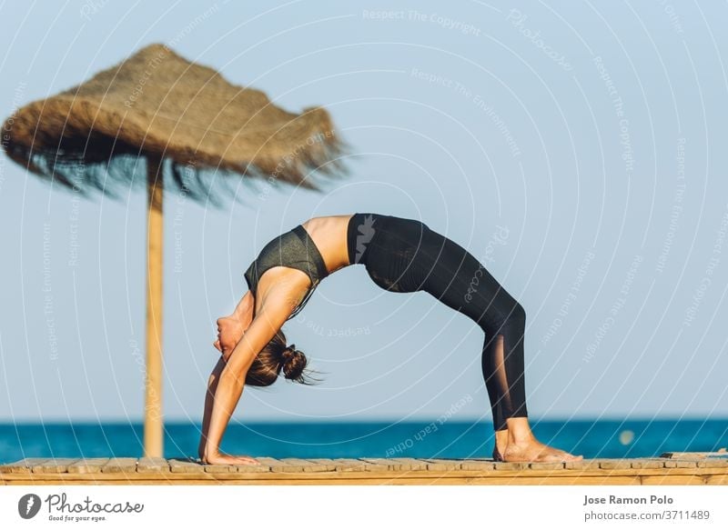 young woman doing yoga bridge exercise on the beach in front of the sea next to umbrella on people meditation person sport fitness relaxation wellness life sand