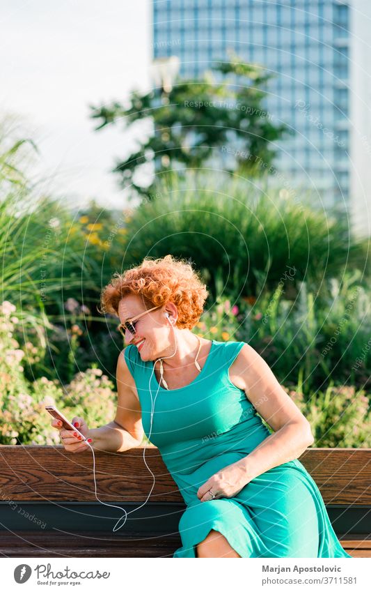 Mature woman listening to the music on her smartphone in the park 60s adult age aged beautiful bench casual caucasian cheerful city connectivity earphones