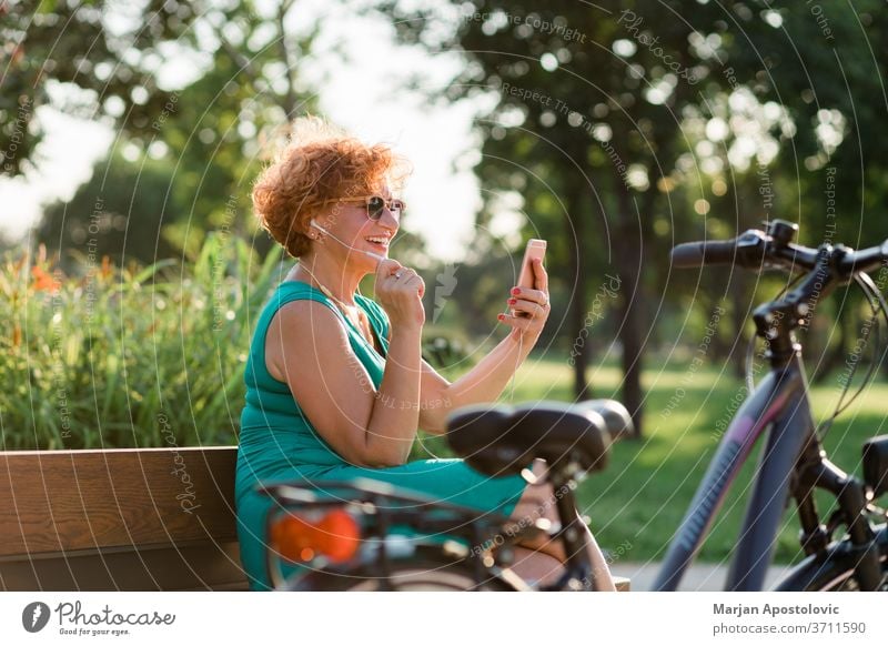 Mature woman using video call on her smartphone in the park 60s age aged app bench bike caucasian cheerful communication connectivity earphones elderly enjoying