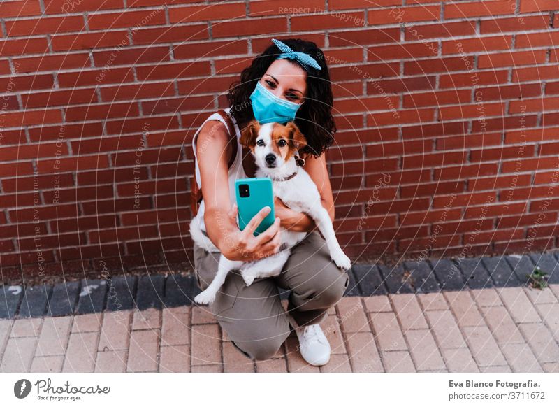young woman walking outdoors wearing protective mask, taking a picture with mobile phone of cute jack russell dog. New normal concept street new normal pet