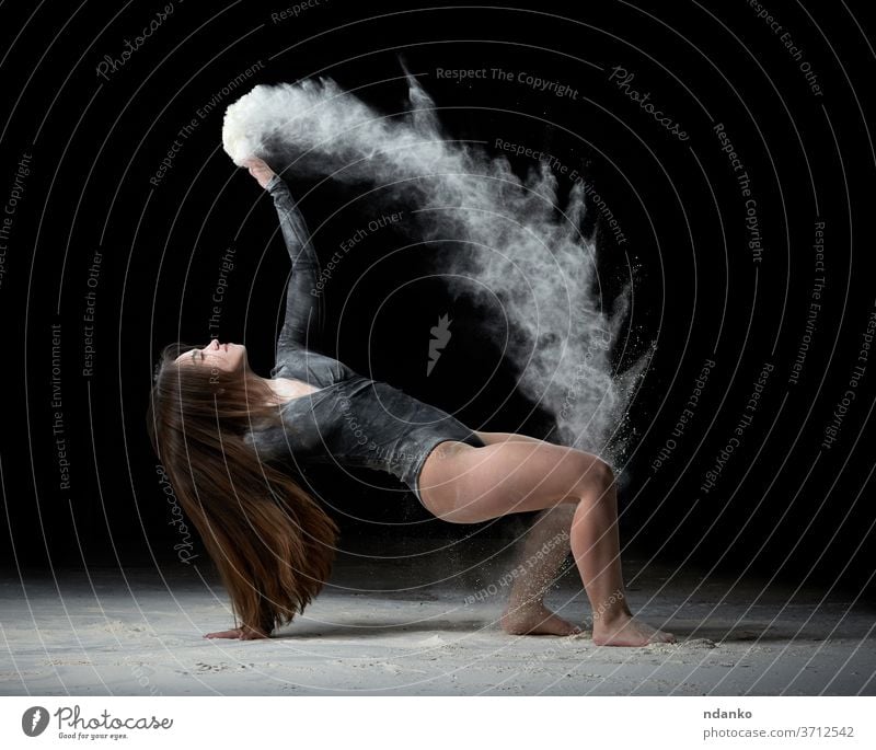 young beautiful woman with long hair is dressed in a sports black bodysuit and sits on the floor and throws white flour over her graceful gymnast up gymnastic