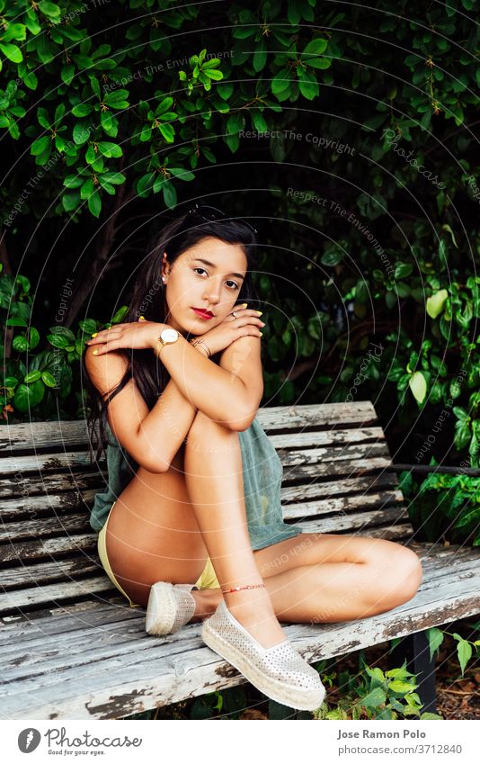 Latin young girl with long black hair and blue dress, with crossed arms sitting on wooden bench among greenery, looking at camera beauty black eyes branches