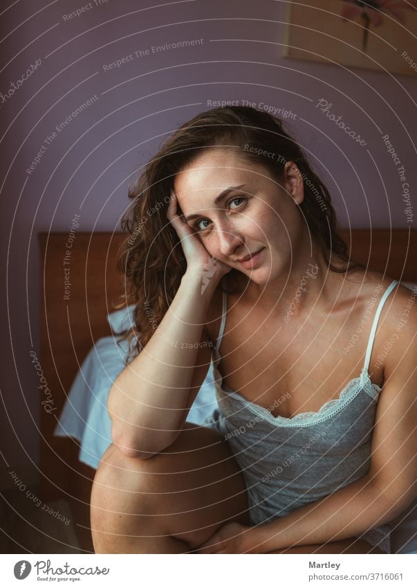 Pleased female in nightwear rumpling hair and looking at the camera while sitting on soft bed in morning woman bedroom indoors relaxation dreaming pillow sexy