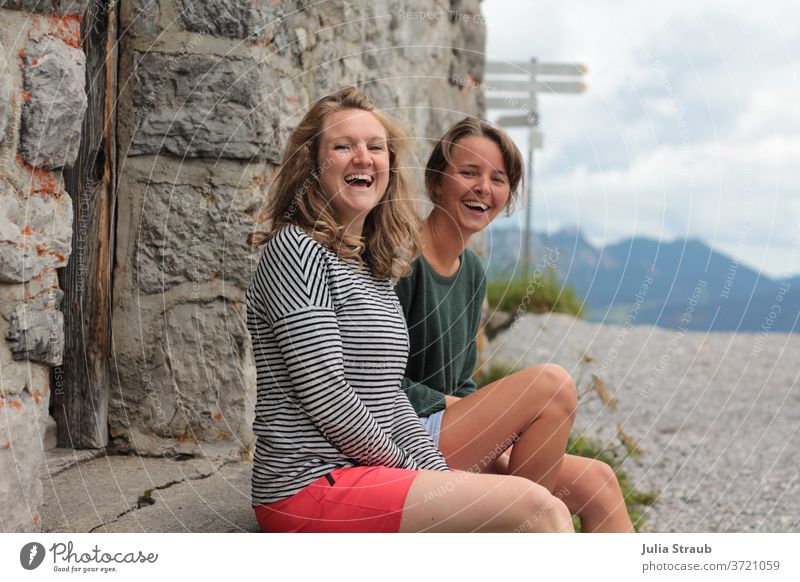 two young ladies are sitting laughing on the step in front of a stone house in the mountains Mountain hike Hiking Break take a break Laughter muck about fun Joy