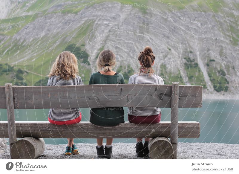 Three young women sitting on a rustic wooden bench at a mountain lake Hiking Break rest tranquillity Mountain Gray Stony Wooden bench Rustic Alpine petrol Lake