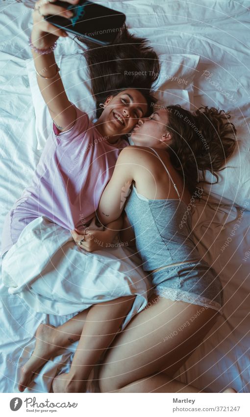 Portrait of two pretty friends taking selfie on cell phone lying in a bed with white sheets, smiling happily. lesbian at home bedroom indoors love woman pillow