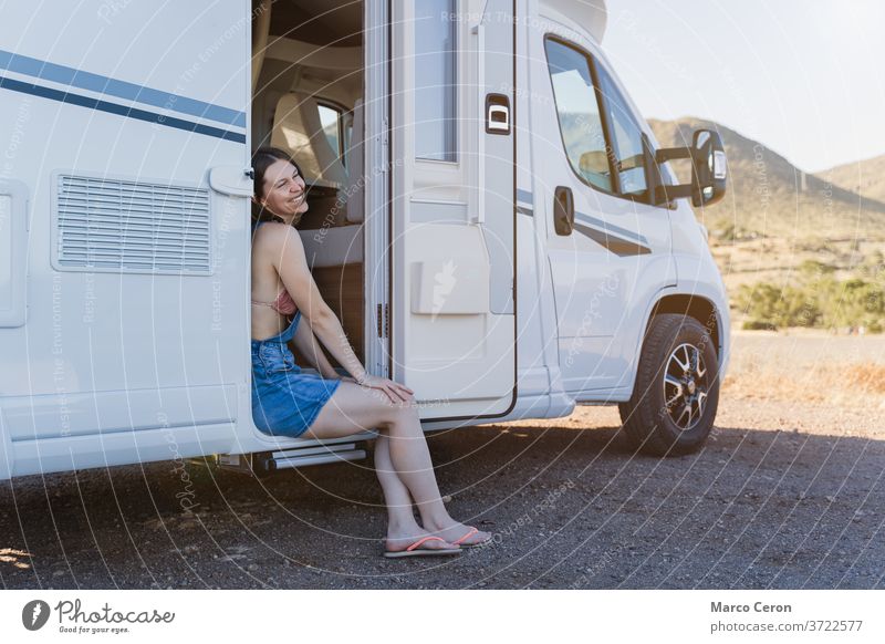 Smiling Young woman sitting onthe door of her motor home parked on desertic nature landscape on a sunny day smiling lifestyles young van mountains exploration