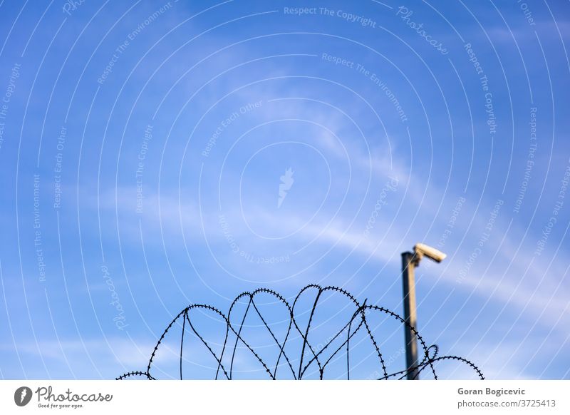 Security camera behind barbed wire fence on the prison wall danger border sky jail camp razor protection security crime surveillance blue metal closeup boundary