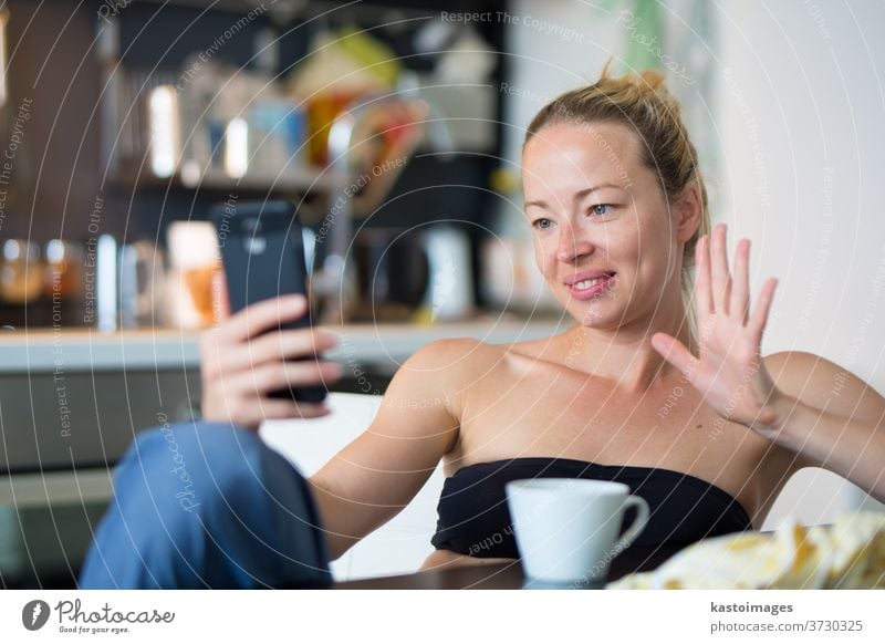 Young smiling cheerful pleased woman indoors at home kitchen using social media on mobile phone for chatting and staying connected with her loved ones. Stay at home, social distancing lifestyle.
