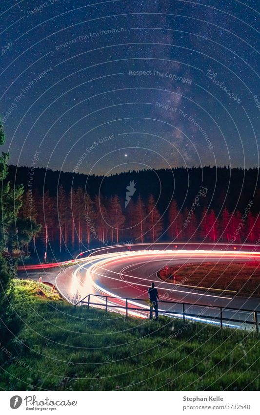 Milkyway, long exposure of a man standing at a railing in front of a curvy street, while cars are passing by and leave cartrail light. milkyway wonderful