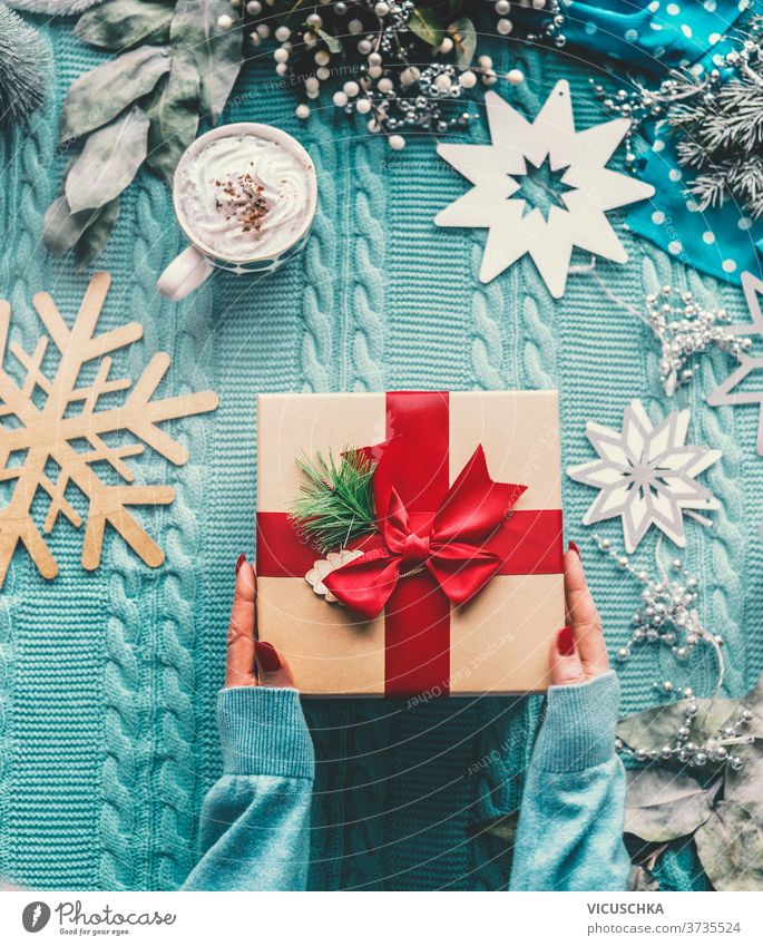 Women hand in pullover holding Christmas gift with red ribbon. Blue knitted blanket with cup of cappuccino. Snowflakes and winter decoration. Top view. Flat lay