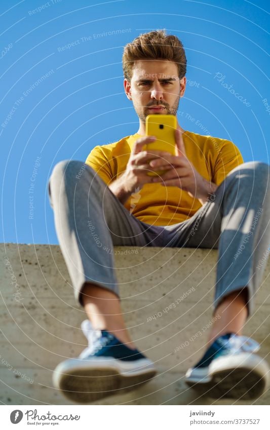 Man using his smartphone sitting on a ledge outside man male lifestyle outdoor young guy hairstyle cell background modern casual communication millennial white