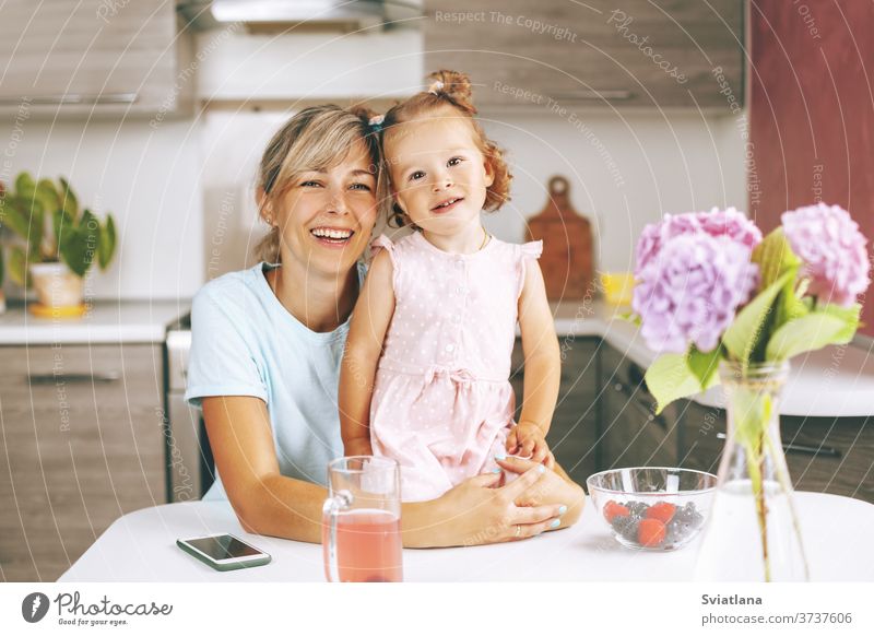 Portrait of an attractive young woman and her little daughter sitting in the kitchen and smiling. On the table is a bouquet of hydrangeas, berries and a glass of juice, side view