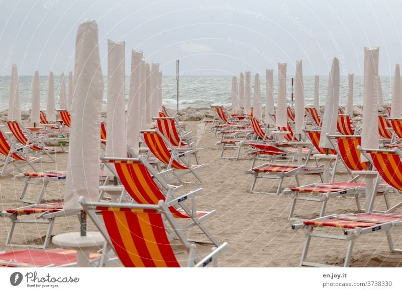 no more summer feeling - folded up deckchairs, parasols and folding chairs on the sandy beach in Italy in front of a sea with waves to the horizon and overcast sky