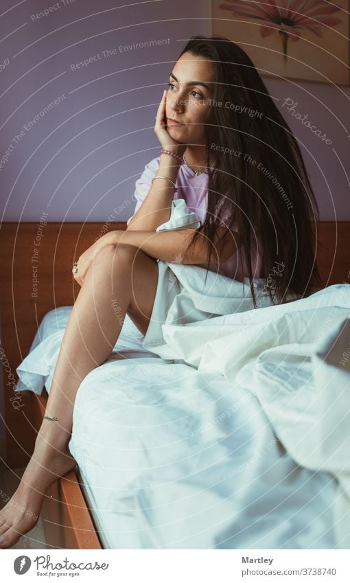 Pleased female in nightwear touching her face and looking at the window while sitting on soft bed in morning. people woman adult one portrait girl room bedroom