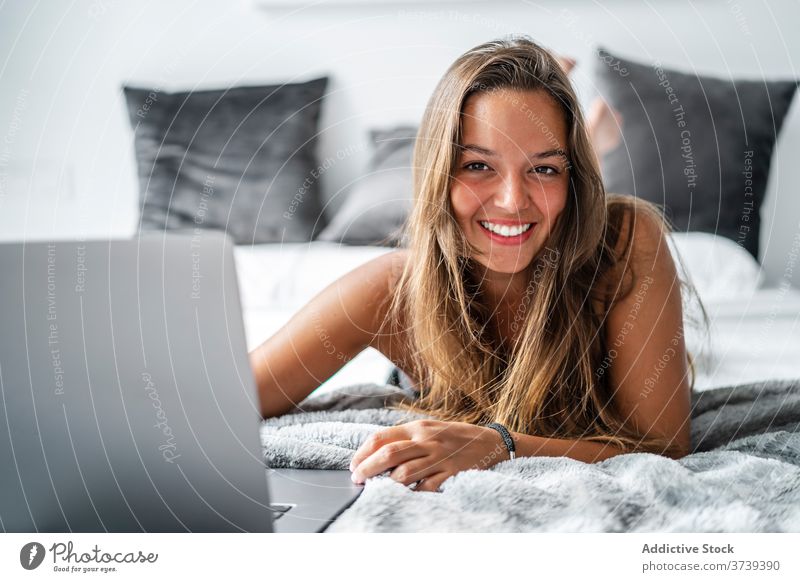 Smiling young woman in sleepwear covering herself with the pillow while  sitting and has one arm by her head on comfortable bed and relaxing in  morning at home. - a Royalty Free