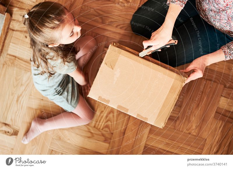 Woman unpacking a cardboard box parcel in room at home. Little girl waiting for opening a gift in package view floor unboxing tape daughter woman filler foam