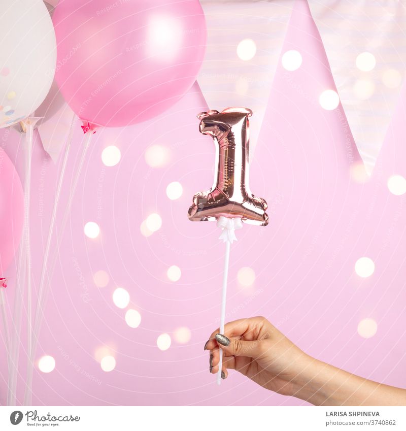 Decoration for one birthday party. Female hand holding golden balloon in form of 1 number on pink background. Sparkles wallpaper decor. celebration celebrate