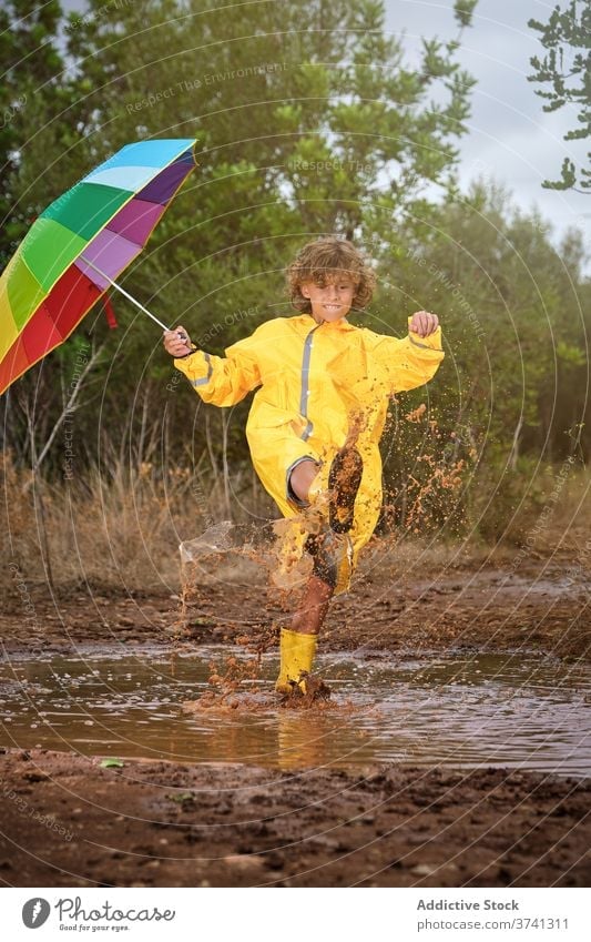 Boy with boots and raincoat holding an umbrella and playing in a puddle joke schoolboy shower attitude rainbow curly gesture splash enjoyment pouring wet