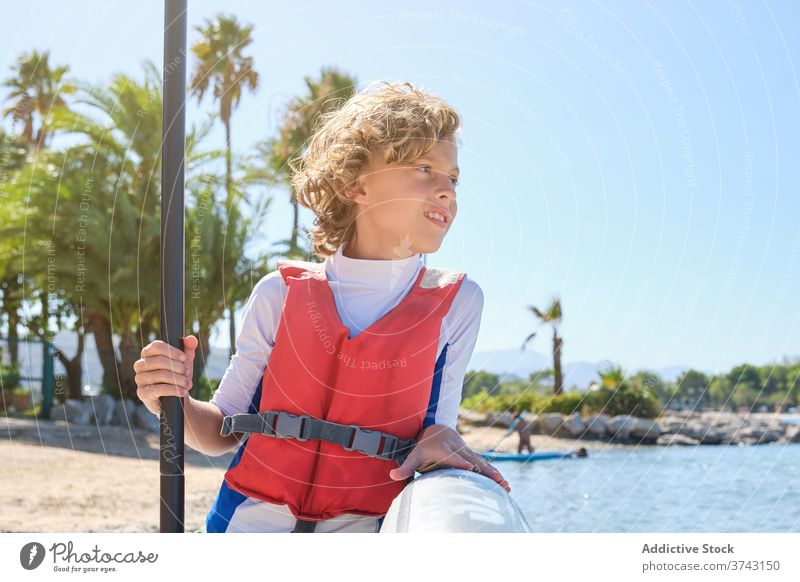 Distracted boy wearing a vest leaning against a paddle surfboard and holding the paddle stick rescue exercise kid outfit surfing tranquility exercising protect