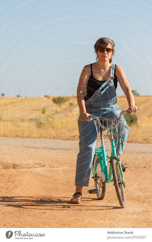 Calm woman on bicycle in rural area road sand sunlight overall bike countryside serene female sunglasses denim summer nature relax casual carefree vacation