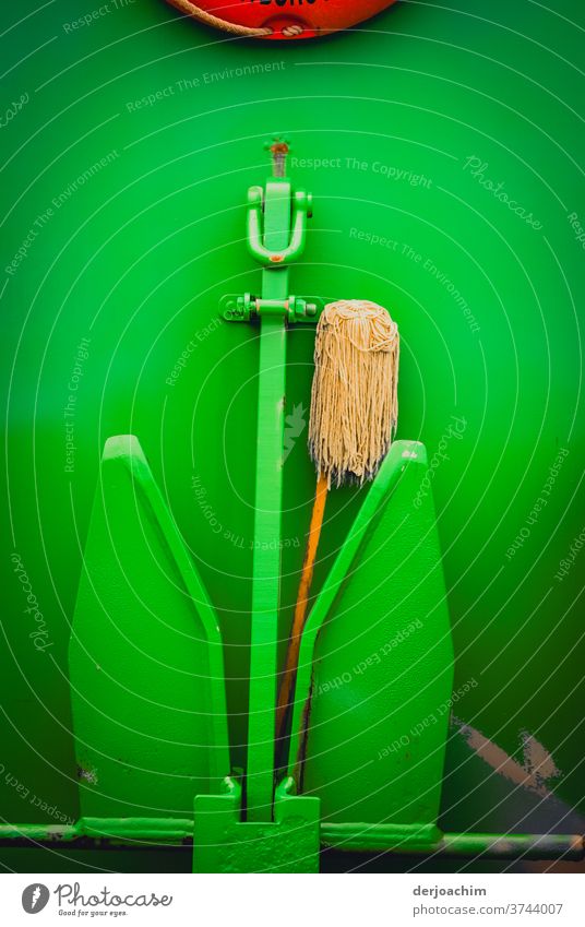 Green painted raised anchor on the railing on a ship. A wiper broom placed in the middle. Also called a feudel. Above them all you can see a red ring, the rescue ring.