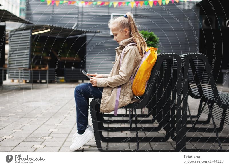 After school, a cute girl sits on a bench waiting for the bus, studies on a tablet online and communicates via video. backpack street schoolgirl learning