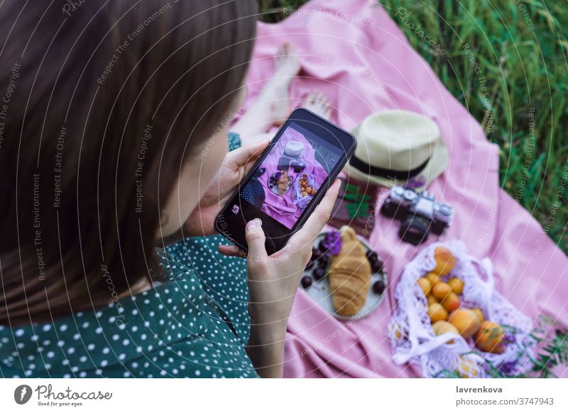 Female taking picture of picnic on green grass outdoors communication smartphone female social media post food blanket pink selfie camera croissant pastry