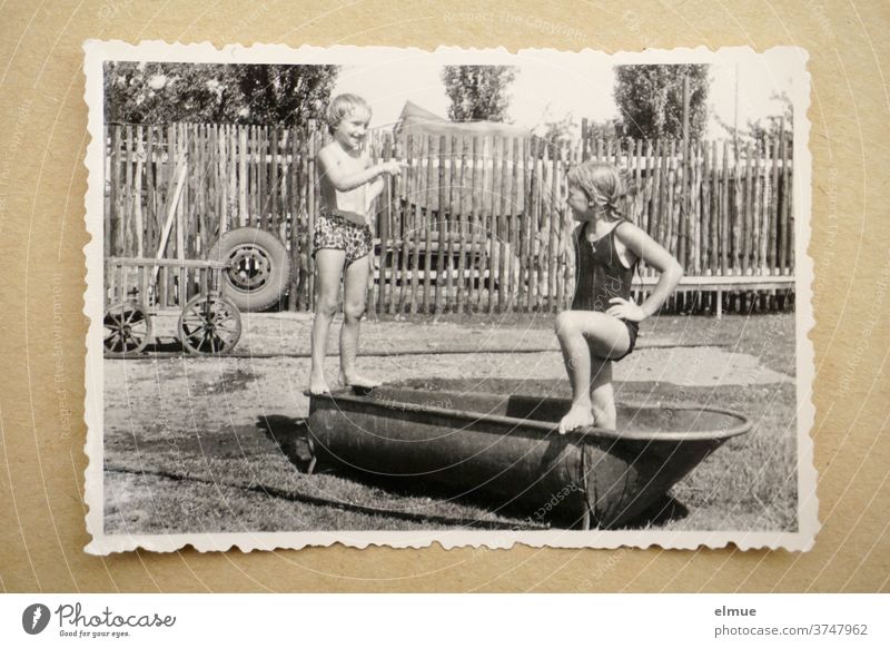 Memories of the 1960s - a black and white photo print with a deckle edge lies on beige paper and shows two girls bathing in an old zinc bathtub in a rural environment with a fence, handcart and car tire