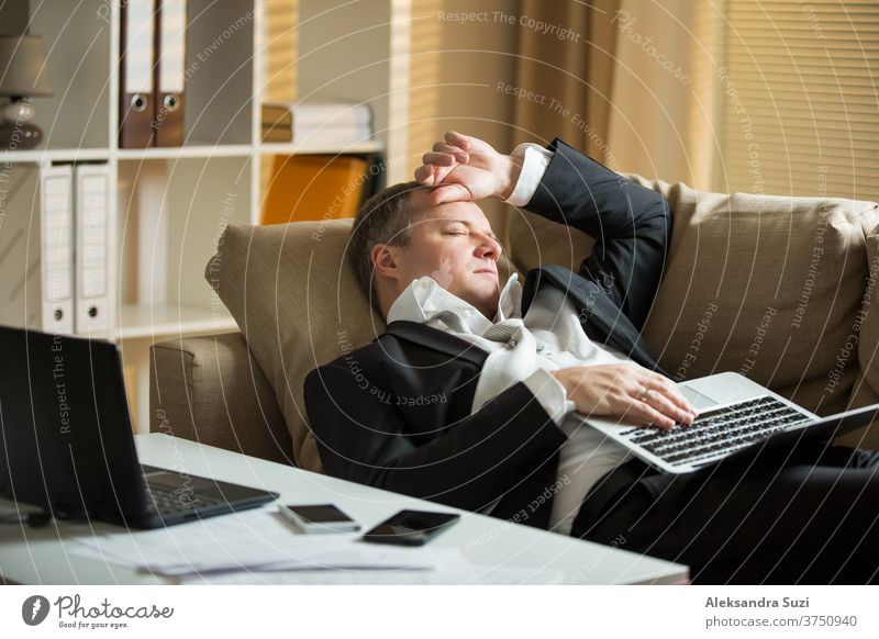 Businessman in suit lying on a couch with two cellphones and laptops, sleeping. Exhausted man relaxing in office early morning. Responsible executive working fell asleep.