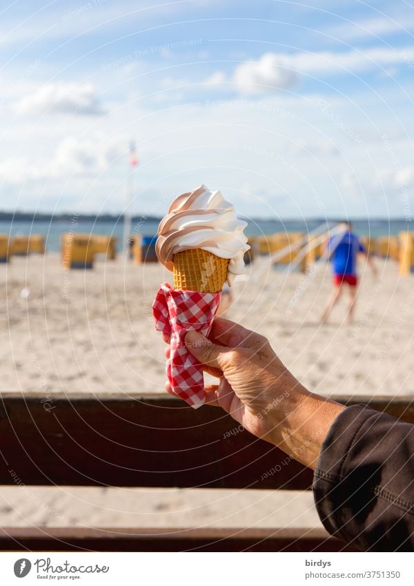 A hand holds an ice cream waffle with soft ice cream. blurred background with beach, beach chairs, sea and a person. nice weather with sunshine. Blue sky, clouds