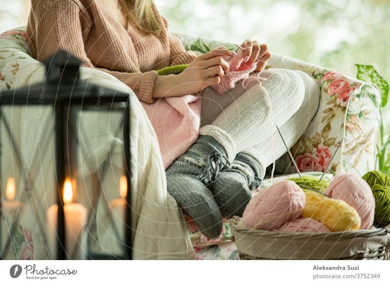 Young woman sitting home in a chair by the window wearing knitted warm sweater, knitting with needles. Cozy room decorated with lanterns and candles. Scenic winter view of pine trees in snow in window