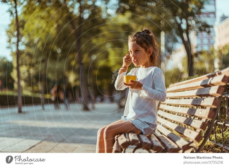 Hispanic kid sitting on a park bench with beautiful trees in the background. She is eating an ice cream. outside real happy sunset season natural seasonal fun