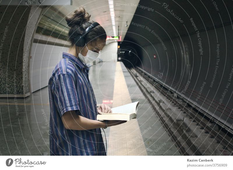 Masked teenager with headphone reading book while waiting for the subway to come metro underground man young student millennial university college adult mask