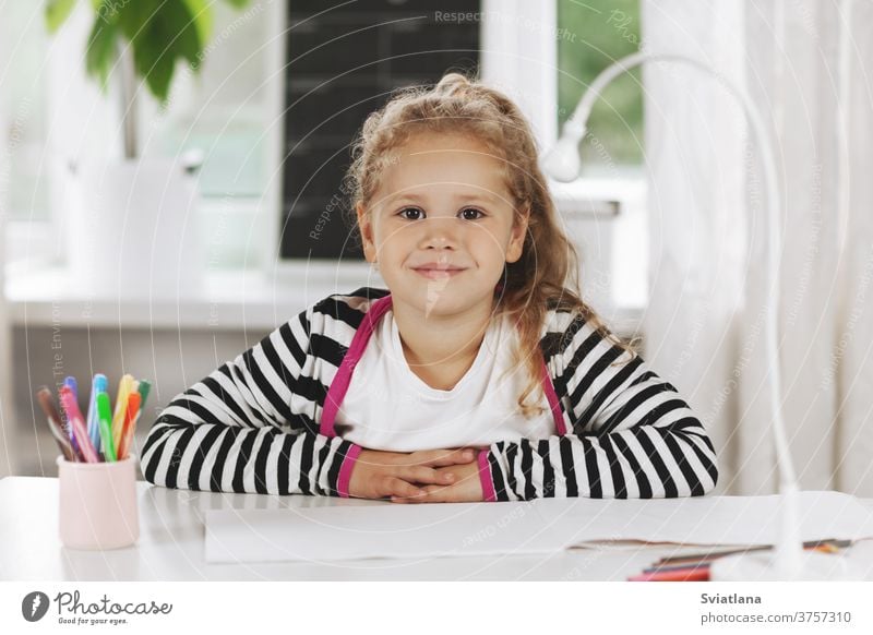 Portrait of a charming little girl sitting at a table. The girl is sitting at the table, on the table is a sketchbook, colored pencils. Homework, home training, social distance