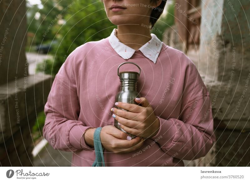 Female in pink sweatshirt holding sustainable metal eco bottle with her hands outdoors, selective focus closeup fingers female woman girl zero waste water