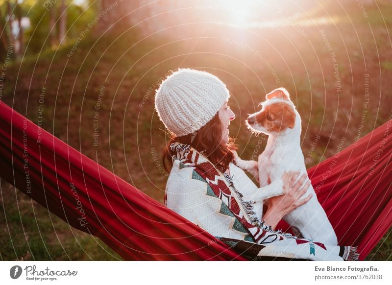 young woman relaxing with her dog in orange hammock. Covering with blanket. Camping outdoors. autumn season at sunset lying hammock jack russell pet nature park