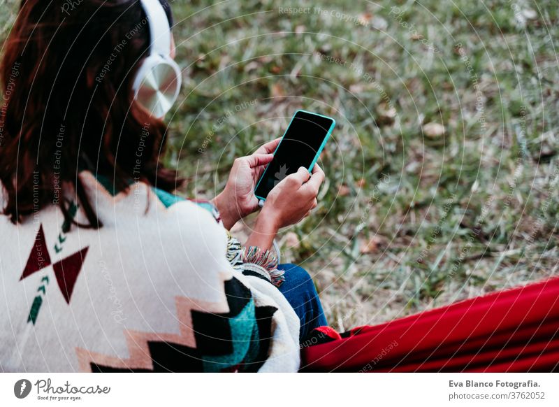 back rear view of woman relaxing in hammock and listening to music on mobile phone and headset. autumn season. camping concept earphones technology internet app