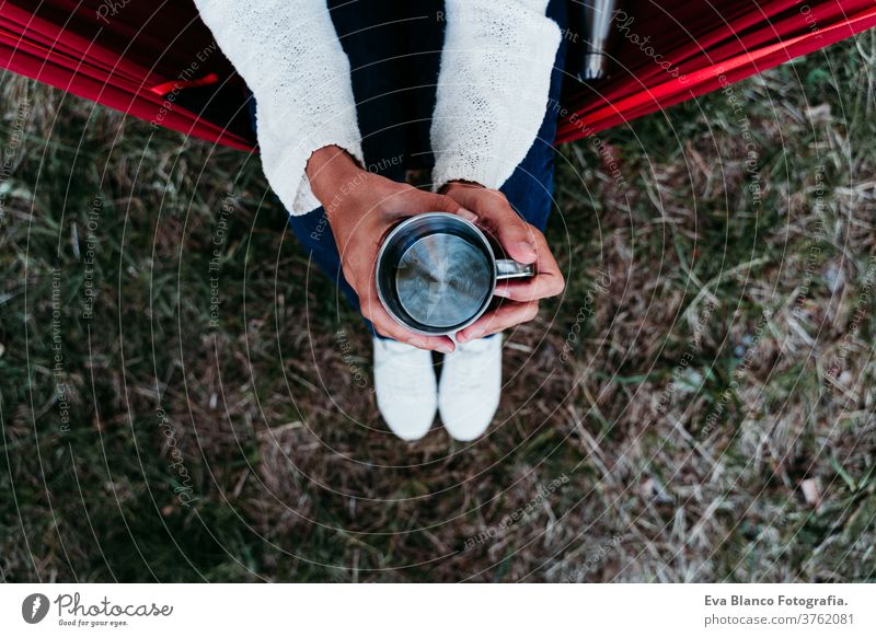 top view of woman holding metallic mug of water, relaxing in hammock at sunset. autumn season thermos unrecognizable feet lying outdoors nature orange park