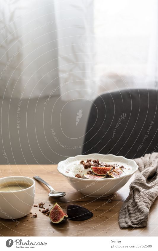 Set breakfast table with granola, yoghurt and fresh figs and a cup of coffee. Window light, morning sun. Breakfast table Yoghurt Fig Coffee Cup bowls Table