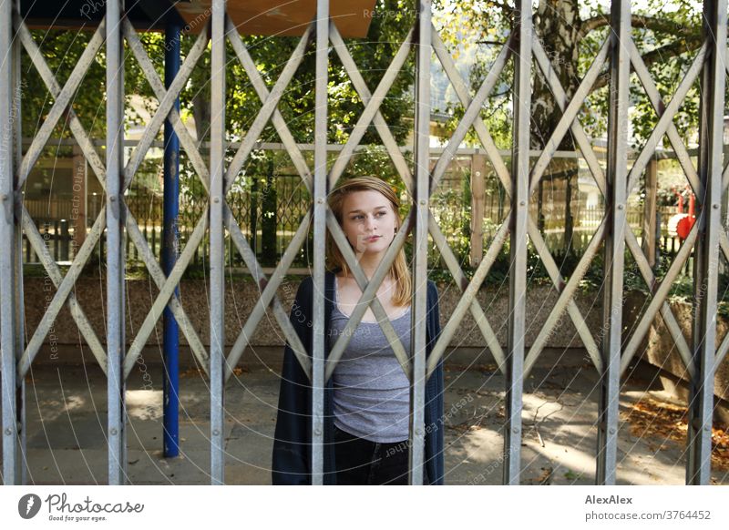 Close portrait of a young woman standing behind a sliding gate and looking through it Woman Young woman Slim already athletic Blonde youthful 18-25 years