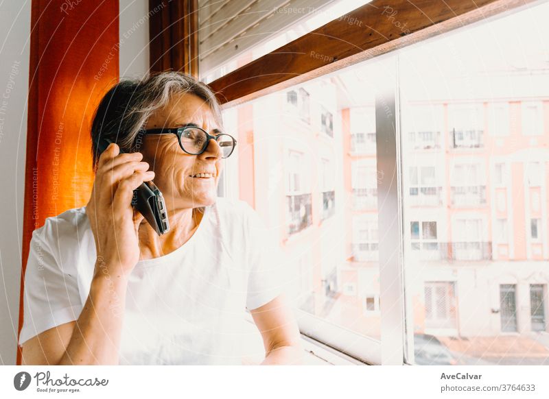 Close up of an old woman making a call on the mobile phone and smiling while looking through the window grandmother indoor retirement person communication