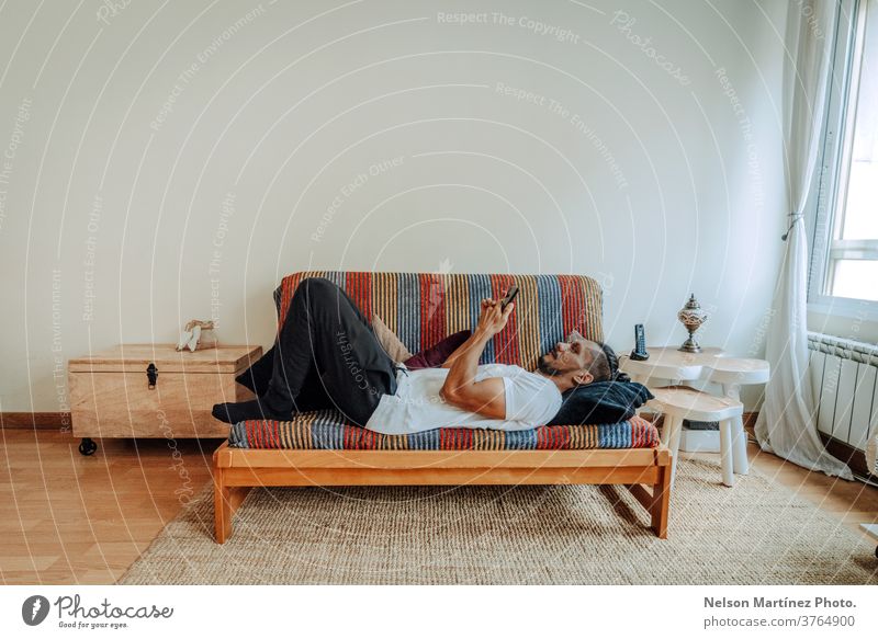Hispanic man lying on a colourful coach relaxed. He is using a smartphone. mobile indoor home mobile phone person people morning 1 working call communication