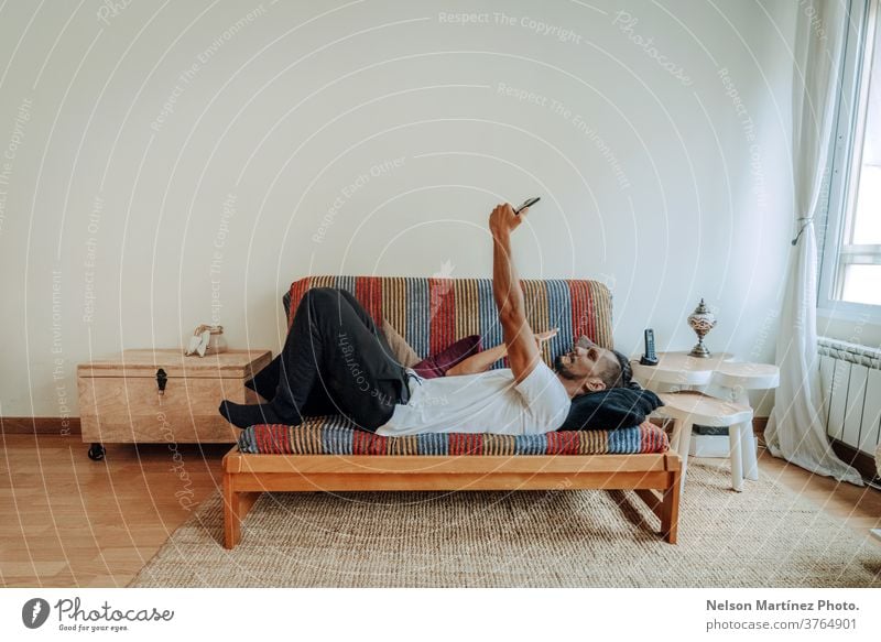 Hispanic man lying on a colourful couch relaxed. He is texting and using a smartphone. mobile indoor home mobile phone person people morning 1 working call