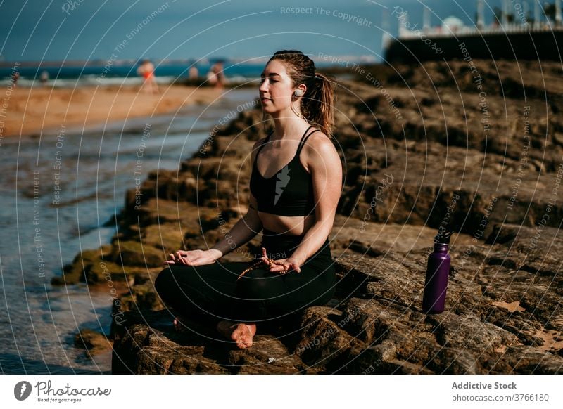 Tranquil woman sitting in Lotus pose and meditating lotus pose meditate yoga listen earphones river shore padmasana zen female stress relief mindfulness stone